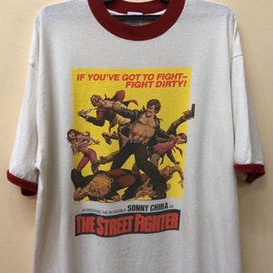 Puede incluir: Camiseta blanca con cuello de canal&eacute; rojo y una impresi&oacute;n de p&oacute;ster de pel&iacute;cula vintage de "The Street Fighter" con Sonny Chiba. El p&oacute;ster tiene el texto "If you've got to fight - fight dirty!"