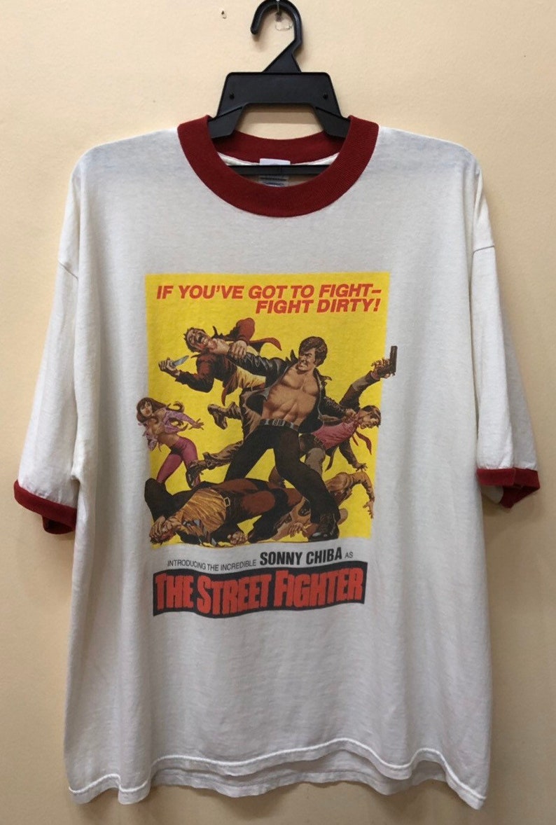 Puede incluir: Camiseta blanca con cuello de canal&eacute; rojo y una impresi&oacute;n de p&oacute;ster de pel&iacute;cula vintage de "The Street Fighter" con Sonny Chiba. El p&oacute;ster tiene el texto "If you've got to fight - fight dirty!"