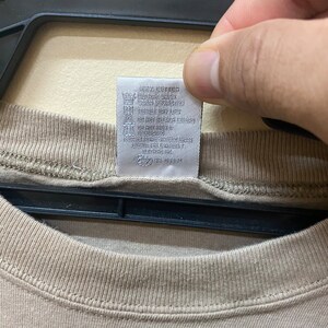 Może przedstawiać: Beżowa koszulka z białą metką pielęgnacyjną z napisem "Only Cotton, Wash Machine Cold, Tumble Dry Low, Do Not Bleach, Do Not Iron, Do Not Dry Clean, Made in USA, RN # 56883".