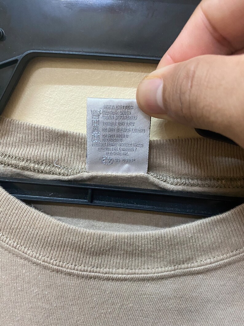 Może przedstawiać: Beżowa koszulka z białą metką pielęgnacyjną z napisem "Only Cotton, Wash Machine Cold, Tumble Dry Low, Do Not Bleach, Do Not Iron, Do Not Dry Clean, Made in USA, RN # 56883".