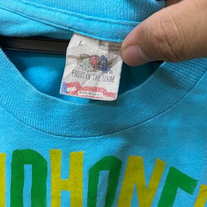 May include: A light blue t-shirt with a yellow, green, and brown graphic print that says "Honey" on it. The shirt has a Fruit of the Loom tag with a size L.