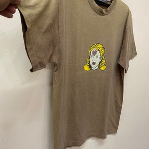 K&ouml;nnte beinhalten: Ein braunes T-Shirt mit einem Aufdruck eines Frauenkopfes mit einem dritten Auge in der Mitte der Stirn. Das dritte Auge ist ein roter Kreis mit dem Wort "Third" darin.