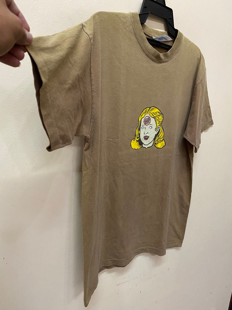 K&ouml;nnte beinhalten: Ein braunes T-Shirt mit einem Aufdruck eines Frauenkopfes mit einem dritten Auge in der Mitte der Stirn. Das dritte Auge ist ein roter Kreis mit dem Wort "Third" darin.