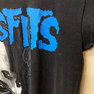 May include: Black t-shirt with a blue "SISIS" graphic and a black and white image of a person with long black hair. The image is of a person's face and shoulders. The t-shirt has a TM logo in the bottom right corner.