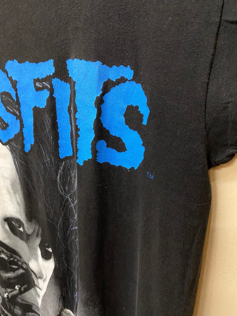May include: Black t-shirt with a blue "SISIS" graphic and a black and white image of a person with long black hair. The image is of a person's face and shoulders. The t-shirt has a TM logo in the bottom right corner.
