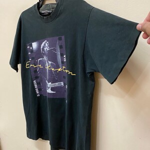 Pu&ograve; includere: Maglietta nera con una foto di Eric Clapton che suona la chitarra. La foto &egrave; in una cornice di pellicola in bianco e nero. Il testo "D 400 DELTA" e "12A" sono visibili sulla pellicola. Il testo "Eric Clapton" &egrave; scritto in corsivo giallo.