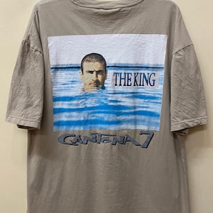 Może przedstawiać: Beżowa koszulka z nadrukiem graficznym przedstawiającym mężczyznę w wodzie z tekstem "THE KING" i "CANTONA 7" na plecach.