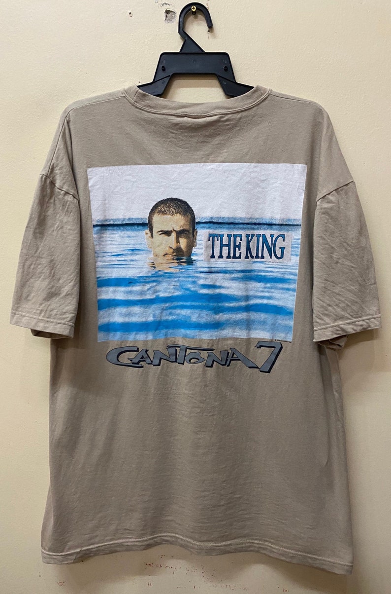 Może przedstawiać: Beżowa koszulka z nadrukiem graficznym przedstawiającym mężczyznę w wodzie z tekstem "THE KING" i "CANTONA 7" na plecach.