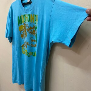 May include: A light blue t-shirt with a graphic print of a band called "Mudhoney" in yellow, green, and brown. The band's name is written in a bold, stylized font.