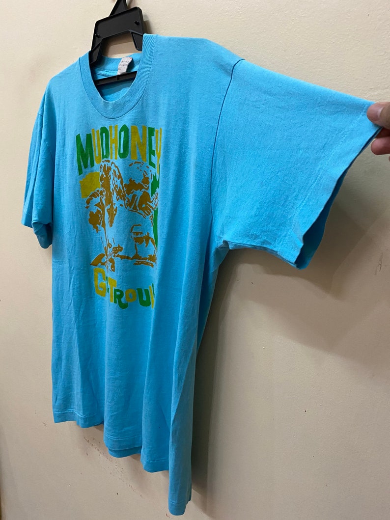 May include: A light blue t-shirt with a graphic print of a band called "Mudhoney" in yellow, green, and brown. The band's name is written in a bold, stylized font.