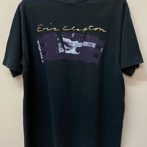 Pu&ograve; includere: Maglietta nera con una stampa dorata della firma di Eric Clapton e una foto della sua mano che suona la chitarra. La foto &egrave; incorniciata da una pellicola.