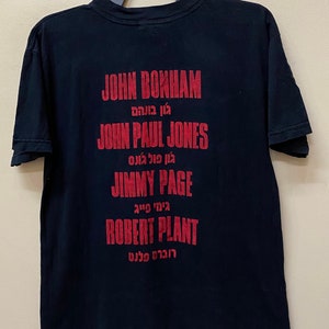Op de afbeelding: Zwart T-shirt met rode tekst die de namen van de bandleden van Led Zeppelin vermeldt: John Bonham, John Paul Jones, Jimmy Page en Robert Plant. De namen zijn in het Engels en Hebreeuws geschreven.