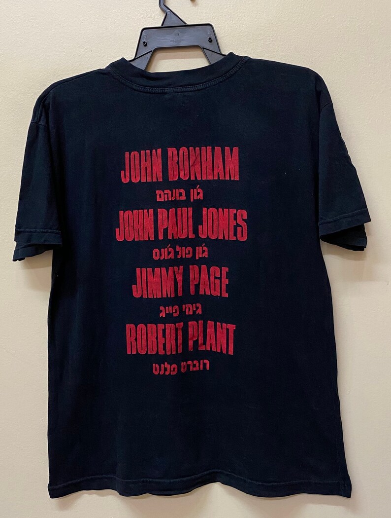 Op de afbeelding: Zwart T-shirt met rode tekst die de namen van de bandleden van Led Zeppelin vermeldt: John Bonham, John Paul Jones, Jimmy Page en Robert Plant. De namen zijn in het Engels en Hebreeuws geschreven.