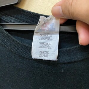 May include: A black t-shirt tag with white text. The tag reads "ADULTOS-L 100% ALGODON HECHO EN MEXICO INSTRUCCIONES AL REVERSO ADULTES-L 100% COTON FABRIQUE AUX MEXICO INSTRUCTIONS AU VERSO ADULT-L 100% COTTON MADE IN MEXICO CARE ON REVERSE"