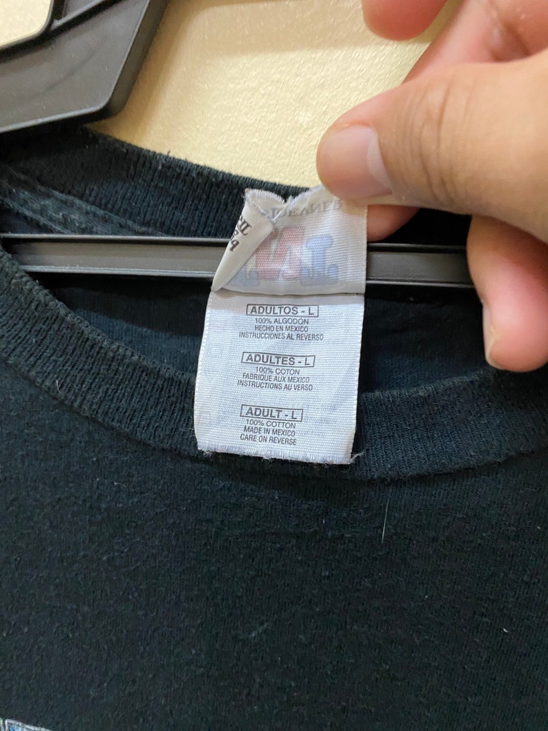 May include: A black t-shirt tag with white text. The tag reads "ADULTOS-L 100% ALGODON HECHO EN MEXICO INSTRUCCIONES AL REVERSO ADULTES-L 100% COTON FABRIQUE AUX MEXICO INSTRUCTIONS AU VERSO ADULT-L 100% COTTON MADE IN MEXICO CARE ON REVERSE"