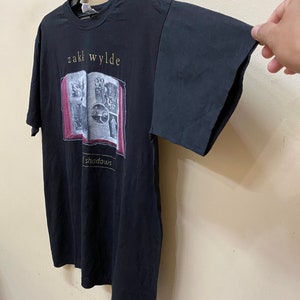 Puede incluir: Camiseta negra con un gr&aacute;fico de un libro con el texto "Zakk Wylde Shadows" impreso en la parte delantera.