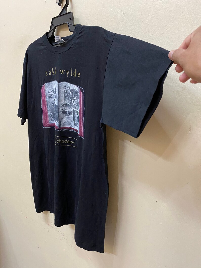 Puede incluir: Camiseta negra con un gr&aacute;fico de un libro con el texto "Zakk Wylde Shadows" impreso en la parte delantera.