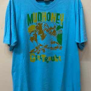 May include: A light blue t-shirt with a graphic print of a band called "Mudhoney" with the words "Girl Trouble" underneath. The band members are depicted in a cartoon style with green, yellow, and brown colors.