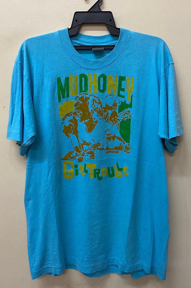 May include: A light blue t-shirt with a graphic print of a band called "Mudhoney" with the words "Girl Trouble" underneath. The band members are depicted in a cartoon style with green, yellow, and brown colors.