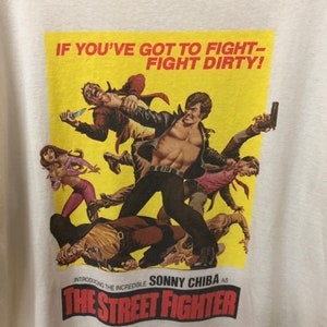 Puede incluir: Una camiseta vintage con un p&oacute;ster amarillo con una escena de la pel&iacute;cula "The Street Fighter". El p&oacute;ster presenta a Sonny Chiba en una escena de pelea con el texto "IF YOU'VE GOT TO FIGHT - FIGHT DIRTY!" y "Introducing the Incredible Sonny Chiba as The Street Fighter".
