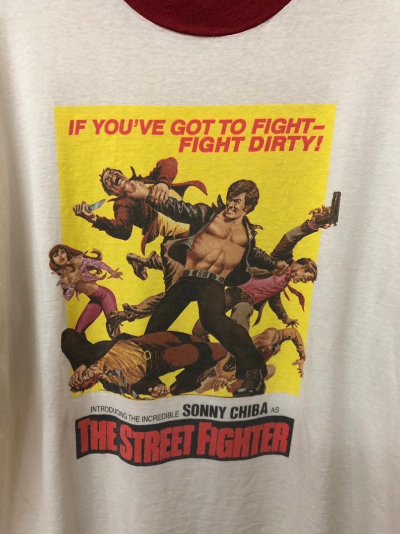 Puede incluir: Una camiseta vintage con un p&oacute;ster amarillo con una escena de la pel&iacute;cula "The Street Fighter". El p&oacute;ster presenta a Sonny Chiba en una escena de pelea con el texto "IF YOU'VE GOT TO FIGHT - FIGHT DIRTY!" y "Introducing the Incredible Sonny Chiba as The Street Fighter".