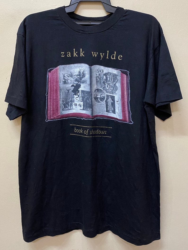 Puede incluir: Camiseta negra con un gr&aacute;fico de un libro abierto con ilustraciones y el texto "zakk wylde book of shadows".