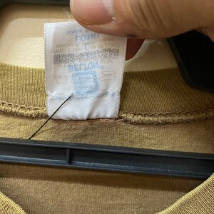 K&ouml;nnte beinhalten: Nahaufnahme eines braunen T-Shirts mit einem wei&szlig;en Etikett, auf dem "The Gap" und "100% Baumwolle" in blauer Schrift stehen.