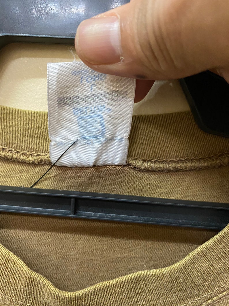 K&ouml;nnte beinhalten: Nahaufnahme eines braunen T-Shirts mit einem wei&szlig;en Etikett, auf dem "The Gap" und "100% Baumwolle" in blauer Schrift stehen.