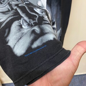 May include: Black long-sleeve t-shirt with a graphic print of a person's face. The text "TM & &copy; 1995 CYCLOPIAN MUSIC INC." is printed on the bottom of the shirt.