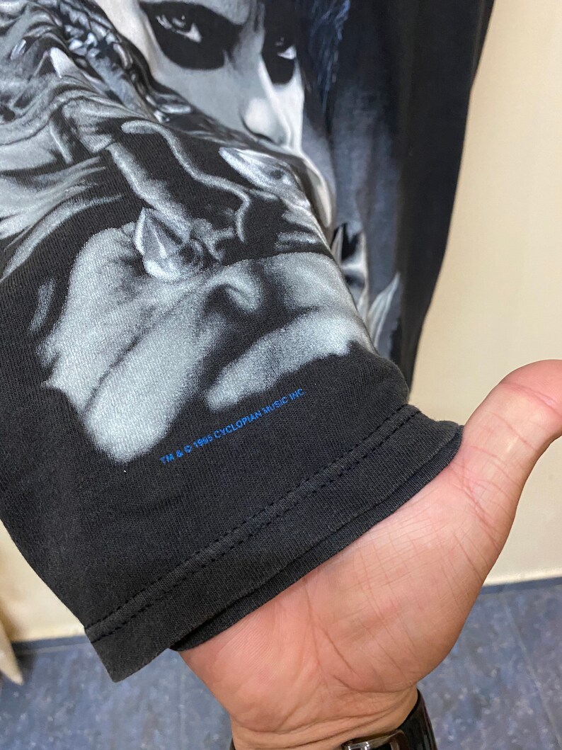 May include: Black long-sleeve t-shirt with a graphic print of a person's face. The text "TM & &copy; 1995 CYCLOPIAN MUSIC INC." is printed on the bottom of the shirt.