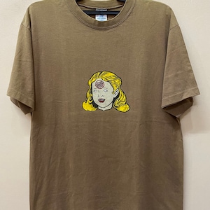 K&ouml;nnte beinhalten: Ein braunes T-Shirt mit einem Aufdruck eines Frauenkopfes mit einem roten, wei&szlig;en und blauen Kreis auf der Stirn. Der Kreis tr&auml;gt die Aufschrift "Santa Cruz".