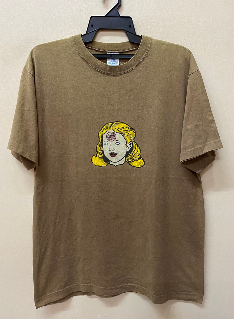 K&ouml;nnte beinhalten: Ein braunes T-Shirt mit einem Aufdruck eines Frauenkopfes mit einem roten, wei&szlig;en und blauen Kreis auf der Stirn. Der Kreis tr&auml;gt die Aufschrift "Santa Cruz".