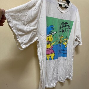 Puede incluir: Camiseta blanca con un estampado gr&aacute;fico verde y azul de un personaje de dibujos animados. El texto "Alice Don't" est&aacute; impreso en el gr&aacute;fico.