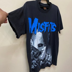 May include: Black t-shirt with a graphic print of a person with black hair and a blue "Misfits" logo.