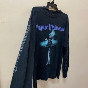 May include: Black long-sleeved t-shirt with a faded image of a person holding a sword. The text "Yngwie Malmsteen" is printed in purple on the front of the shirt. The text "The Seventh Sign World Tour" is printed on the right sleeve.