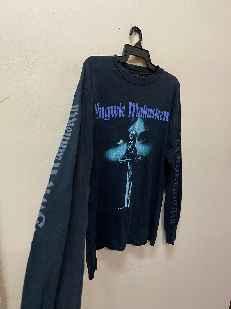 May include: Black long-sleeved t-shirt with a faded image of a person holding a sword. The text "Yngwie Malmsteen" is printed in purple on the front of the shirt. The text "The Seventh Sign World Tour" is printed on the right sleeve.