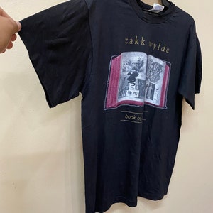 Puede incluir: Camiseta negra con un gr&aacute;fico de un libro con el texto "Zakk Wylde Book of Shadows" impreso en &eacute;l.