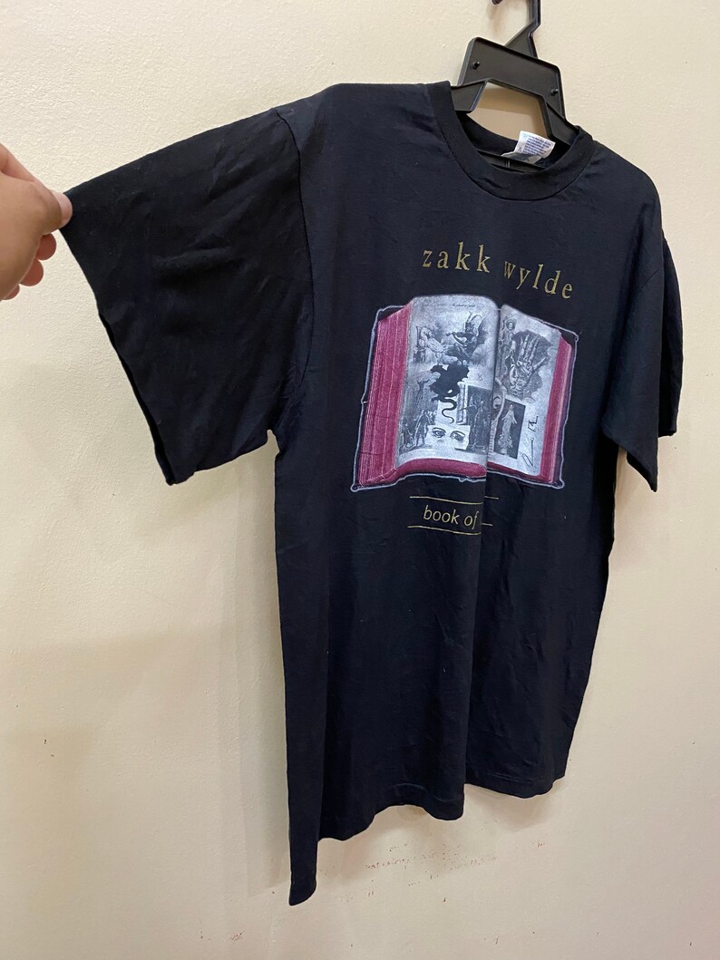 Puede incluir: Camiseta negra con un gr&aacute;fico de un libro con el texto "Zakk Wylde Book of Shadows" impreso en &eacute;l.