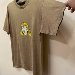 K&ouml;nnte beinhalten: Ein braunes T-Shirt mit einem Aufdruck einer Frau mit einem roten Kreis auf der Stirn. Die Frau hat blonde Haare und tr&auml;gt ein wei&szlig;es Hemd.