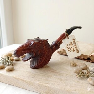 May include: A dark brown wooden tobacco pipe carved into the shape of a dragon head with a black stem. A small rectangular wooden tag is attached to the stem with twine, featuring the text "TWO HEARTS ONE LOVE" and a double heart icon.