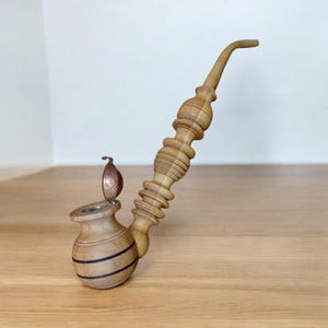 Puede incluir: Una pipa de tabaco de madera clara con un tallo curvado y un cuenco con dos bandas oscuras. El tallo tiene un diseño decorativo tallado y una tapa metálica con bisagras. La madera es de color marrón claro.
