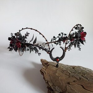 Peut inclure: Couronne florale noire avec des roses rouges et un charme de croissant de lune. La couronne est faite de fil de fer et a une finition noire. Le charme de croissant de lune a une pierre pr&eacute;cieuse rouge au centre.
