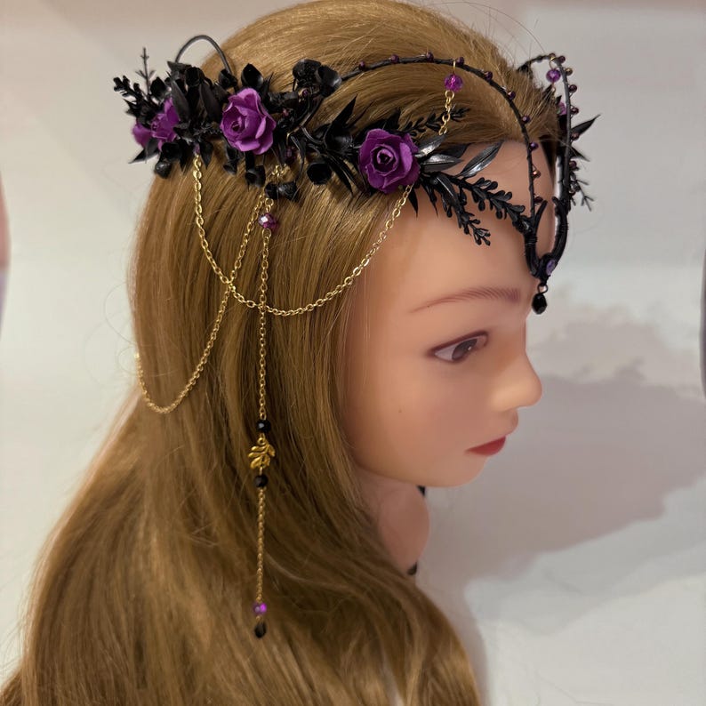 May include: A headpiece featuring black foliage and purple roses arranged along a dark frame. The accessory includes delicate gold-toned chains that drape down the side, accented with small purple beads and a gold leaf charm hanging from the chains.