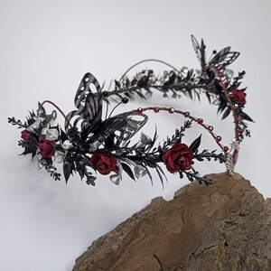Pu&ograve; includere: Una corona floreale nera e rossa con farfalle e dettagli argentati. La corona &egrave; realizzata in filo metallico e ha un design delicato e fantasioso.