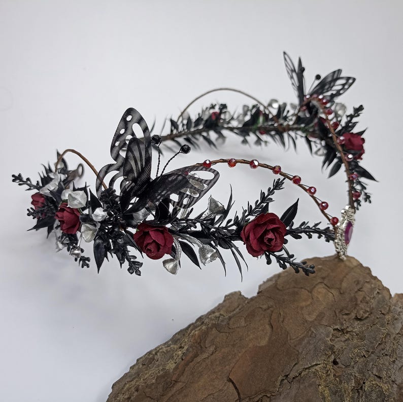 Pu&ograve; includere: Una corona floreale nera e rossa con farfalle e dettagli argentati. La corona &egrave; realizzata in filo metallico e ha un design delicato e fantasioso.
