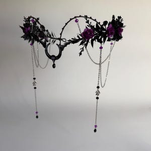 May include: A headpiece featuring a black crescent moon charm at the center, adorned with dark purple roses, black foliage, and silver-toned chains. The design includes small purple and black beads hanging from the chains, creating a layered effect.