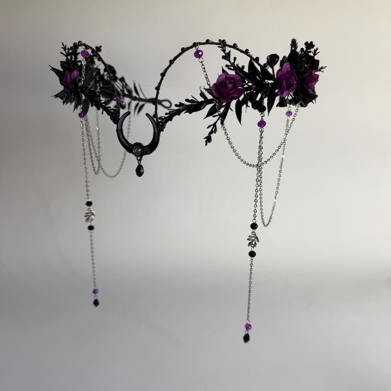 May include: A headpiece featuring a black crescent moon charm at the center, adorned with dark purple roses, black foliage, and silver-toned chains. The design includes small purple and black beads hanging from the chains, creating a layered effect.
