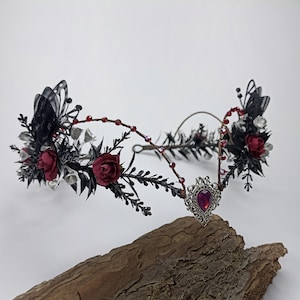 Pu&ograve; includere: Una corona floreale nera e argentata con rose rosse e un ciondolo argentato con una pietra preziosa rossa. La corona &egrave; realizzata in filo metallico ed &egrave; decorata con piume nere e perline argentate.