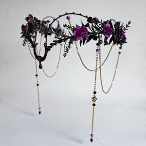 May include: A headpiece featuring a black wire frame adorned with dark purple roses, black leaves, and small purple beads. The design includes delicate gold-toned chains that drape downwards, accented with black and purple beads and a crescent moon charm.