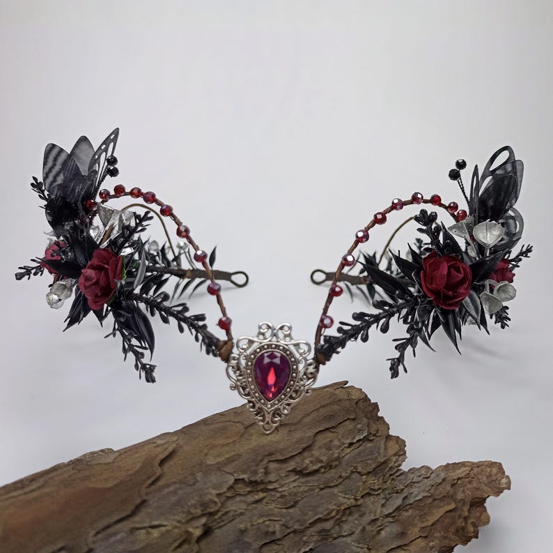 Pu&ograve; includere: Una corona floreale nera e argentata con rose rosse e una grande pietra preziosa rosa scuro al centro. La corona &egrave; realizzata in filo metallico e ha un design delicato e fantasioso.